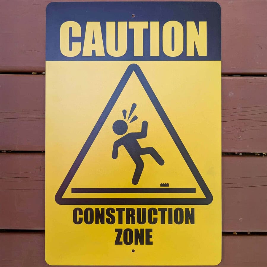 Yellow Caution Construction Zone Metal Sign, front view on wood background, featuring a figure stepping on a brick, by True North Bricks, available at Infinite Brick
